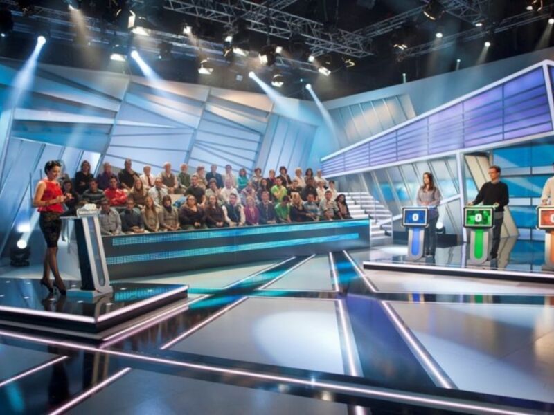 Un panel de concursantes y un presentador en un escenario de televisión con luces y pantallas digitales.
