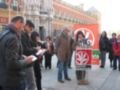 Una persona se encuentra en un podio con una pancarta que dice "Partido Andalucista". La persona está usando un micrófono y parece estar dando una conferencia. Algunos asistentes están observando la persona en el podio, mientras que otros están escribiendo. En el fondo, hay un edificio histórico con ventanas y balcones.