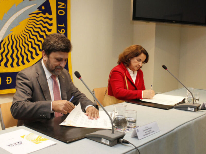 Dos personas sentadas en una mesa, uno de ellos firma un documento mientras la otra toma notas.