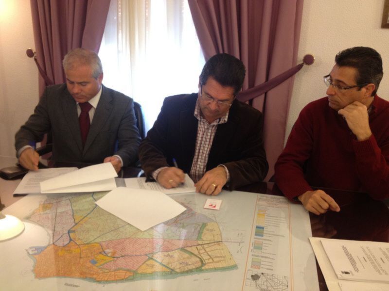 Tres personas sentadas en una mesa, uno de ellos firma un documento mientras los otros observan.