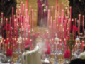 Un sacerdote en vestido blanco bendice a una mesa de altar con velas rojas y flores. En el fondo, se ve un altar dorado con una cruz y una figura en rojo.