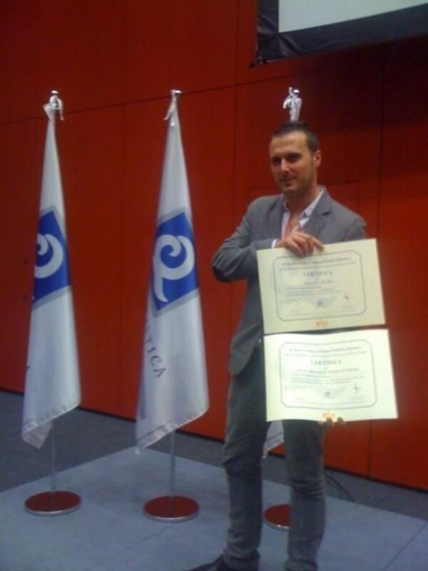 Persona recibiendo un certificado en un evento formal con banderas y pantalla de proyección en el fondo.