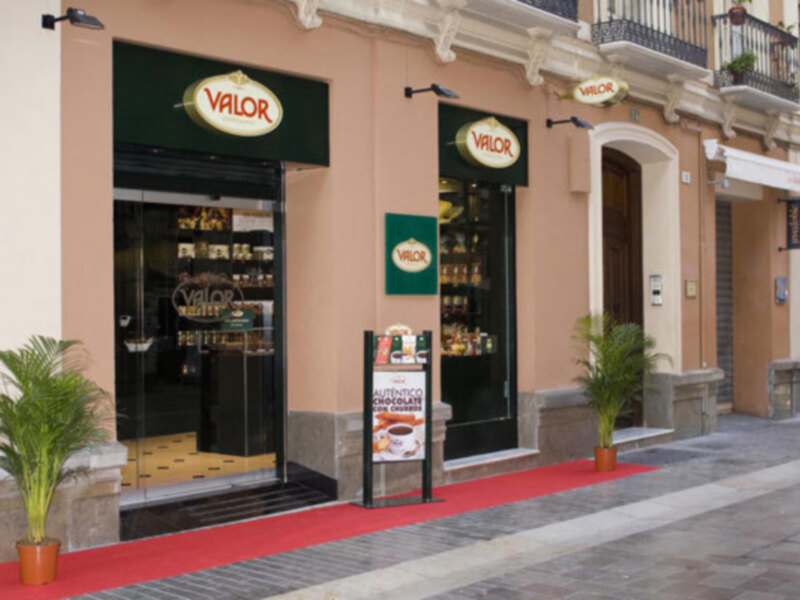 Puerta de entrada de un local comercial con una puerta de cristal y una pancarta de bienvenida en el exterior. La fachada es de color beige y hay plantas en el exterior.