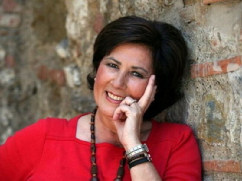 Retrato de una mujer sonriente en rojo, apoyada contra una pared de ladrillo antiguo.