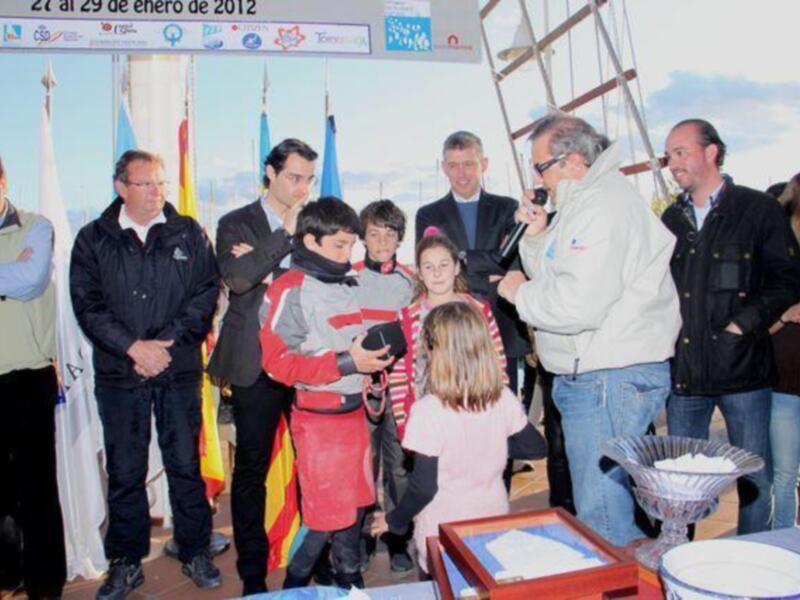 Una familia en un evento público, con una persona en el centro sosteniendo a un niño y otros adultos alrededor, frente a una bandera nacional y un cartel informativo sobre fechas.