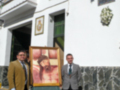 Dos hombres en trajes formales posan frente a una fachada blanca con detalles decorativos y una imagen de Cristo en una capa dorada.