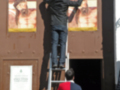 Dos hombres trabajando en una escalera, uno pintando un cuadro y otro observando. La imagen muestra una pared con varios cuadros y un cartel de entrada a la sala de exposiciones.