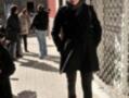 Una mujer camina por una calle, vestida con un abrigo negro, pantalones de mezclilla y botas de invierno, con una campana de la tienda Katia en el fondo.