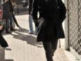 Una mujer camina por una calle, vestida con un abrigo negro y botas marrones. La imagen muestra detalles de su look, incluyendo una chaqueta negra y botas marrones.