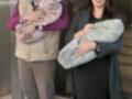Una pareja con una persona embarazada y dos bebés recién nacidos. La imagen muestra a la mujer con un vestido largo y el hombre con una chaqueta de cuadros. El ambiente parece ser urbano, posiblemente en una estación o entrada de edificio.