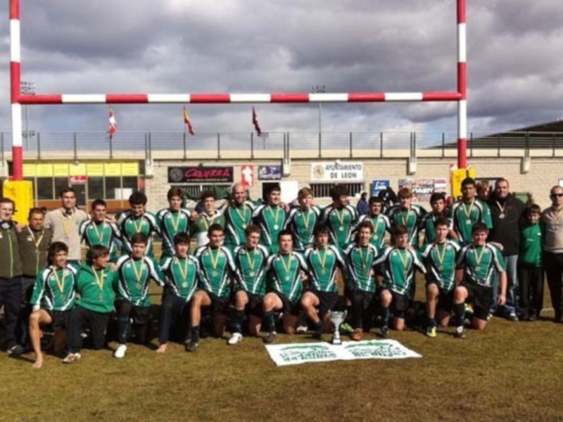Equipo de rugby celebrando su victoria con medallas y trofeo en un estadio con banderas y arco de rugby en el fondo.