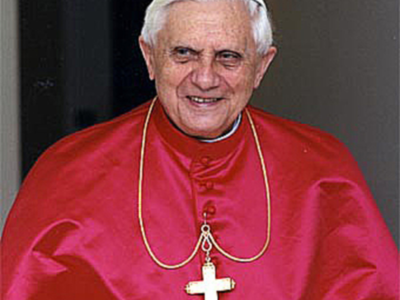 Imponente imagen del Papa Benedicto XVI en su vestimenta papal roja, con una sonrisa y una cruz de oro al cuello.