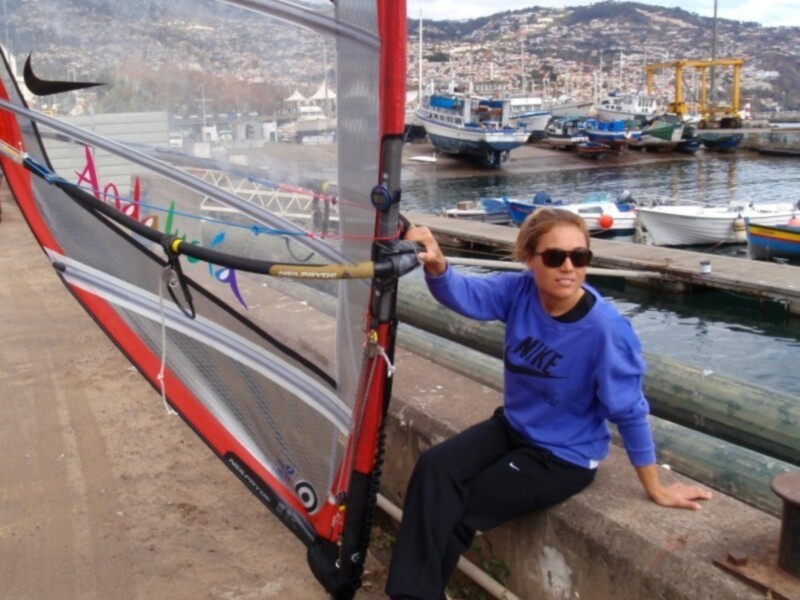 Una persona posa con un vela de windsurf en un muelle, con el mar y barcos al fondo.