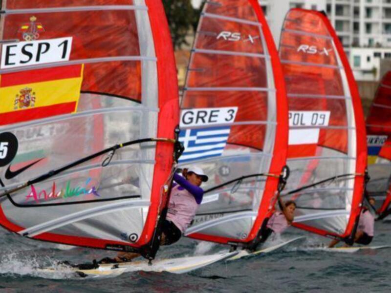 Competidores en acción durante una regata de vela de viento, destacando banderas españolas y griegas.