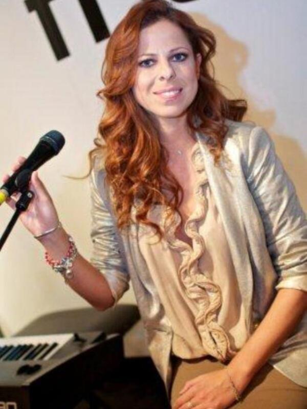 Mujer con cabello largo y rizado sosteniendo un micrófono, posando en un evento con un piano en el fondo.