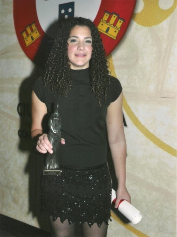 Persona con cabello rizado largo sosteniendo un trofeo y un diploma, posando frente a un escudo con castillos.