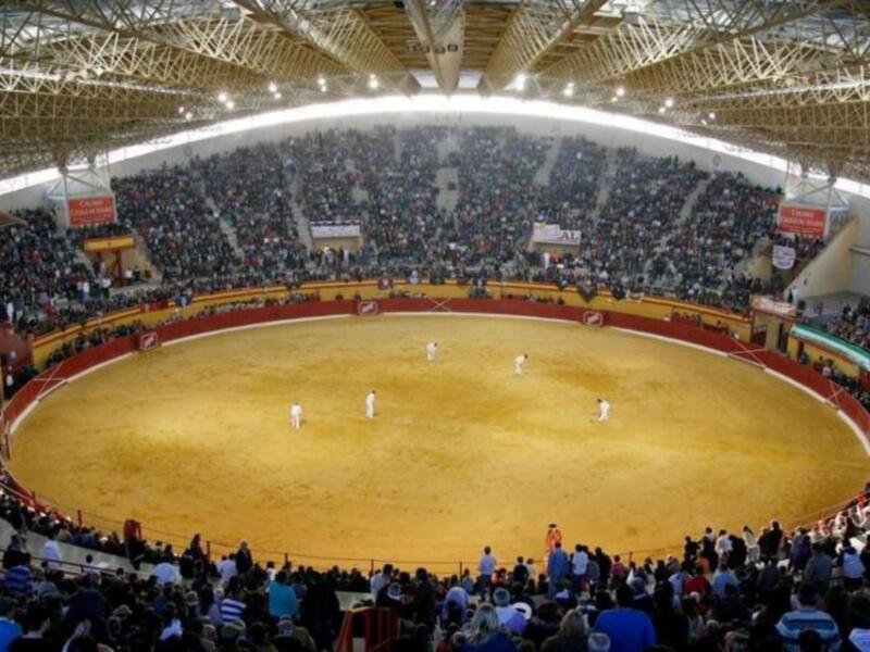 Interior de una arena de toros con un público numeroso y toreros en el centro del ruedo. La estructura tiene una iluminación brillante y una gran capacidad para espectadores.