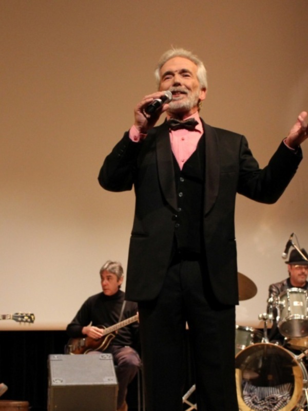 Cantante en traje negro y corbata roja actuando en un escenario con músicos en el fondo.