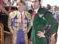 Dos toreros posan juntos en un ambiente festivo. El primero lleva traje de color azul con detalles dorados, mientras que el segundo usa un traje verde brillante con bordados. Ambos son sonrientes y parecen estar en un evento de toros.