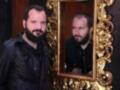 Un hombre con barba mira su reflejo en un espejo dorado.