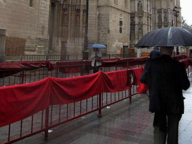 Persona caminando bajo la lluvia con paraguas, cerca de una estructura antigua cubierta con un velo rojo.