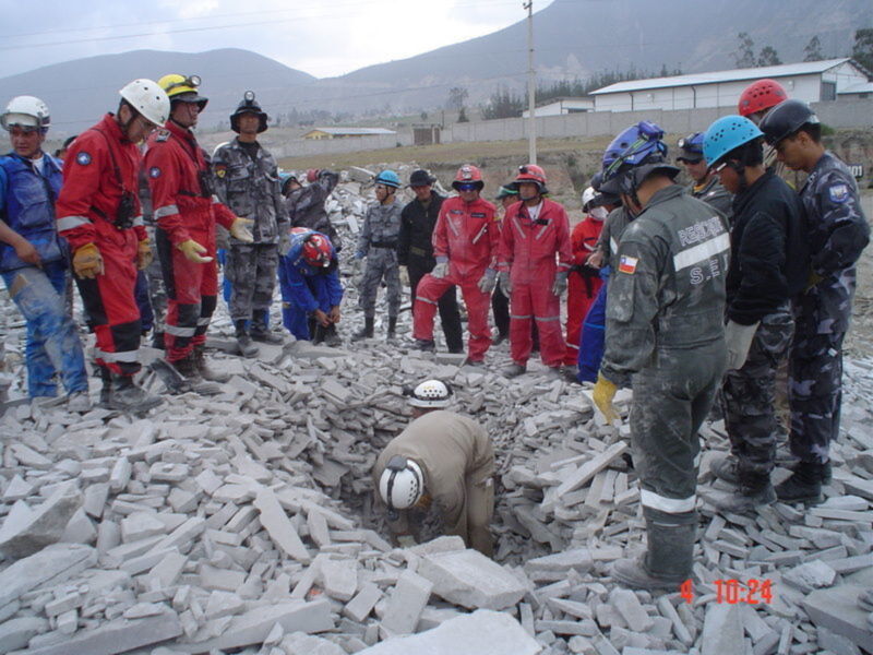Equipo de rescate y supervisión en terreno de desastre, con equipo de protección y supervisando estructuras dañadas.