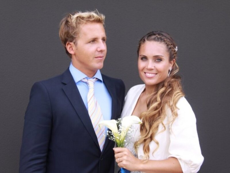 Una pareja joven sonríe mientras el hombre lleva un traje formal y la mujer sostiene una ramita de flores. El fondo es un muro de color gris.