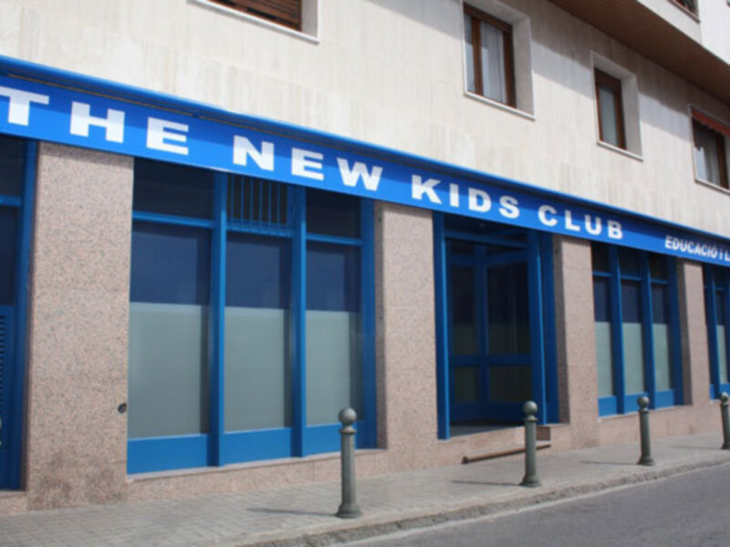 La fachada de un edificio con una tienda llamada 'The New Kids Club' en letras azules y blancas. El edificio tiene ventanas grandes y azules, y una puerta principal. La calle está vacía y hay pequeños postes de metal en el frente del edificio.