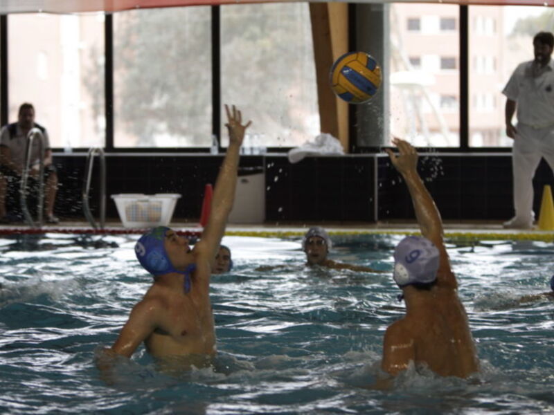 Jugadores de waterpolo en acción en una piscina interior, con un balón en movimiento y varios jugadores en el agua.