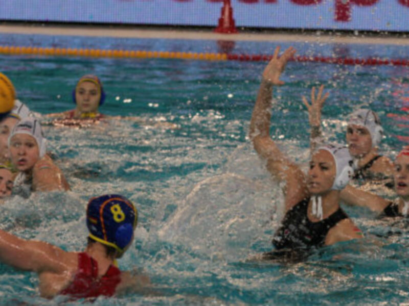 Juego de waterpolo en una piscina con jugadoras en acción. La pelota está en movimiento y varias jugadoras están en la acción de golpearla. La piscina está llenada de agua y hay una línea de natación visible en el fondo.