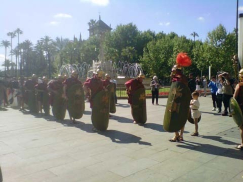 Recreación histórica con soldados romanos en una plaza con fuente y árboles.