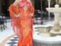 Una mujer con un vestido de flamenco rojo y naranja, con detalles en la cintura y mangas largas. Fondo de una plaza con un fuente, sillas y columnas.