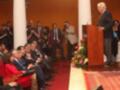 El presidente está dando un discurso en un auditorio lleno de personas. La imagen muestra a varios espectadores, algunos tomando fotos con sus cámaras digitales. El presidente está en un podio con una floración de flores en el frente, y la audiencia está sentada en filas.