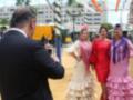 Una persona toma una foto de tres mujeres vestidas con trajes de flamenca en un ambiente festivo. La escena está ambientada en una plaza con edificios y decoraciones al fondo, indicando un evento cultural o festivo.