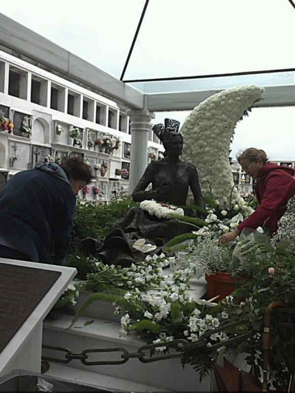 Día de los Muertos en un cementerio, con una estatua adornada con flores y dos personas depositando ofrendas.