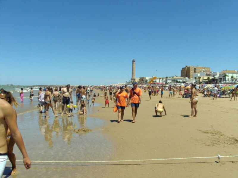 Playa de Muelle Uno, Cádiz, con visitantes disfrutando del sol y el mar bajo un cielo azul despejado.