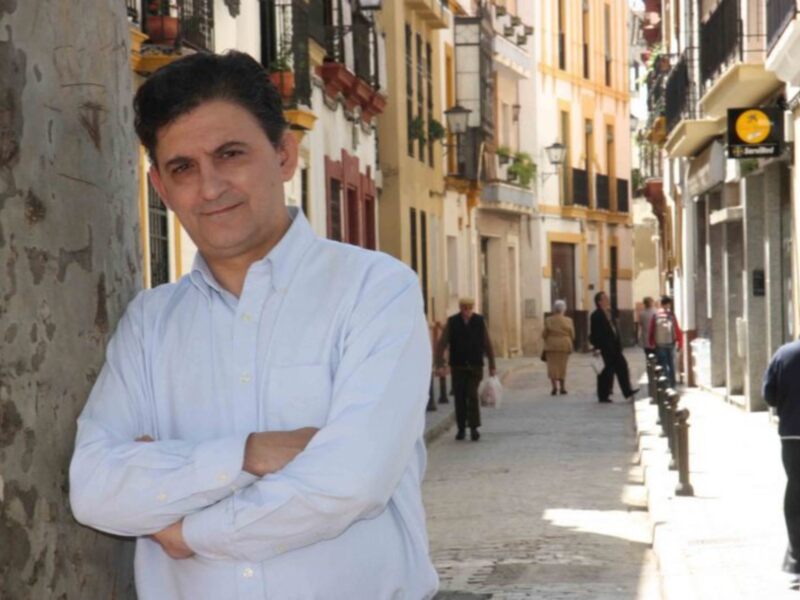Un hombre con camisa azul posa en la calle, rodeado de edificios amarillos y verdes. Detrás de él, se pueden ver otros ciudadanos caminando y una calle con árboles.