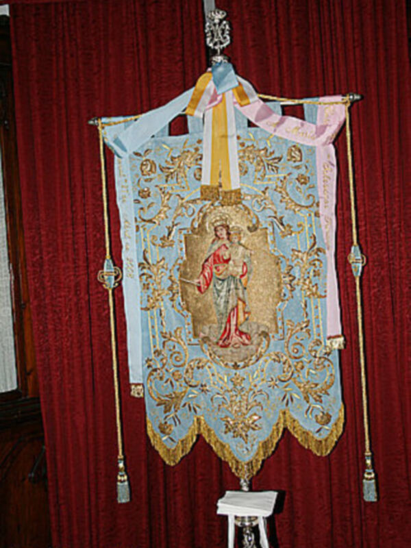 Banderín religioso con bordados y figura central en un soporte ceremonial, colgado en un ambiente con cortina roja.