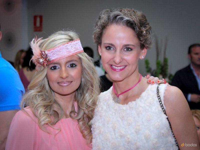Dos mujeres sonrientes, una con cabello largo y rubio con un tocado rosa y la otra con cabello corto y rizado. Ambas están vestidas formalmente, una en rosa y la otra en blanco con detalles de flores. La escena parece ser en un evento formal o fiesta, con otros asistentes en el fondo.
