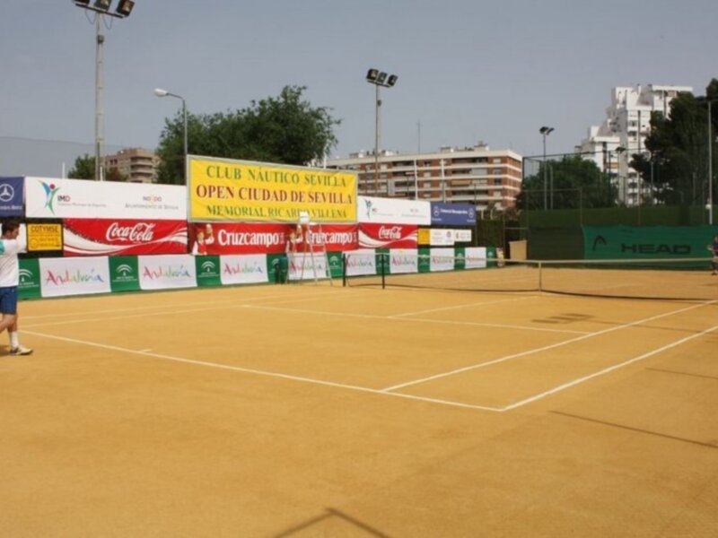 Juego de tenis en un campo de tierra batida con anuncios y banderas de patrocinadores en el Club Náutico Sevilla.