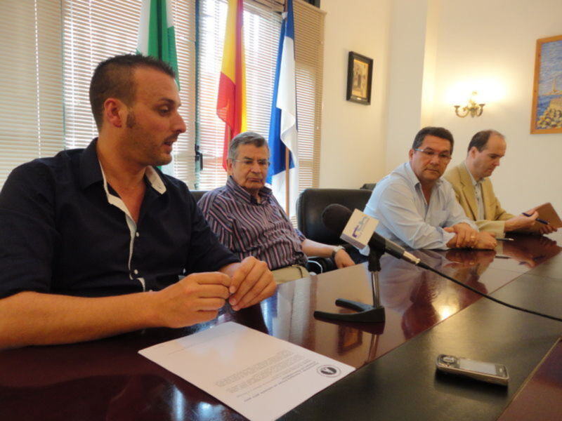 Conferencia de prensa con tres hombres sentados en una mesa, uno hablando en un microfono. Banderas en el fondo.