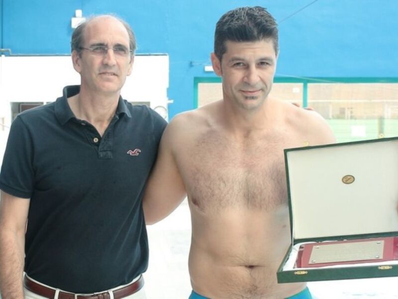 Un hombre con una medalla de oro en una piscina, posando junto a otro hombre vestido con una camisa de polo azul.