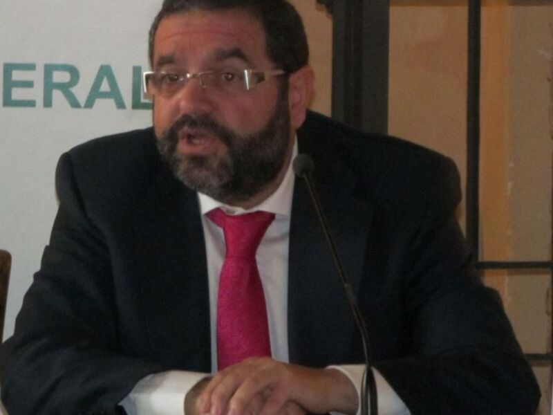 Un hombre con barba y gafas hablando en un evento formal.