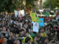 Una multitud de personas en una manifestación, con carteles y banderas, en un ambiente urbano con árboles al fondo.