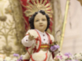 Un niño vestido con una túnica blanca y roja, sosteniendo un palo de mando. La imagen está rodeada por flores y adornos religiosos, en un ambiente festivo.