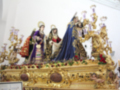 Escena religiosa con tres figuras vestidas en trajes tradicionales, posiblemente de la Virgen María y santos, en un altar dorado con flores y candelabros.
