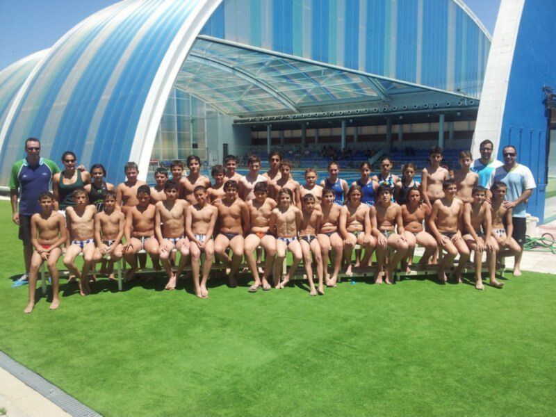 Equipo de natación posando en la piscina cubierta con el estadio al fondo.