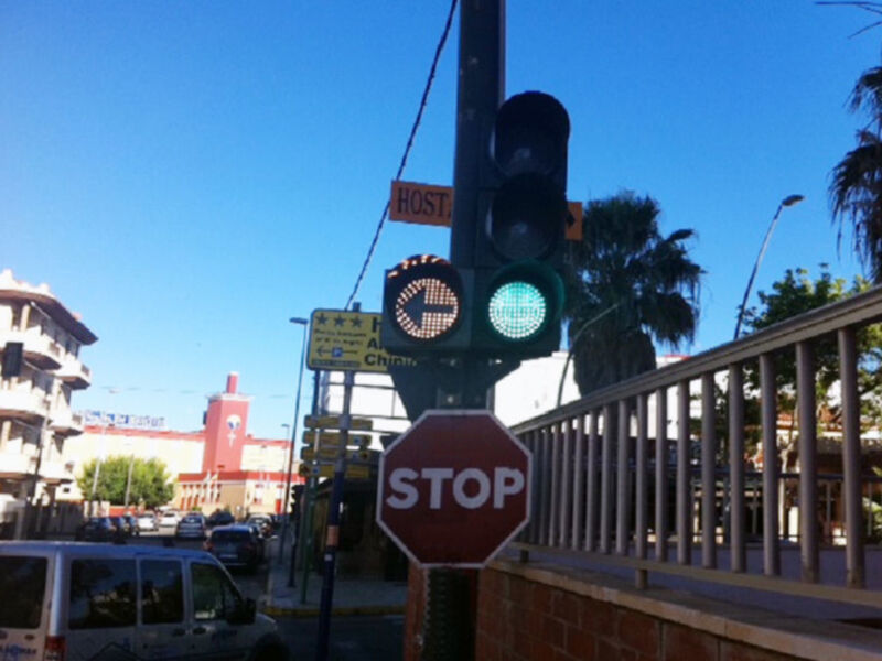 Señal de tráfico con semáforo verde y signo de detención en una calle urbana con edificios y árboles al fondo.