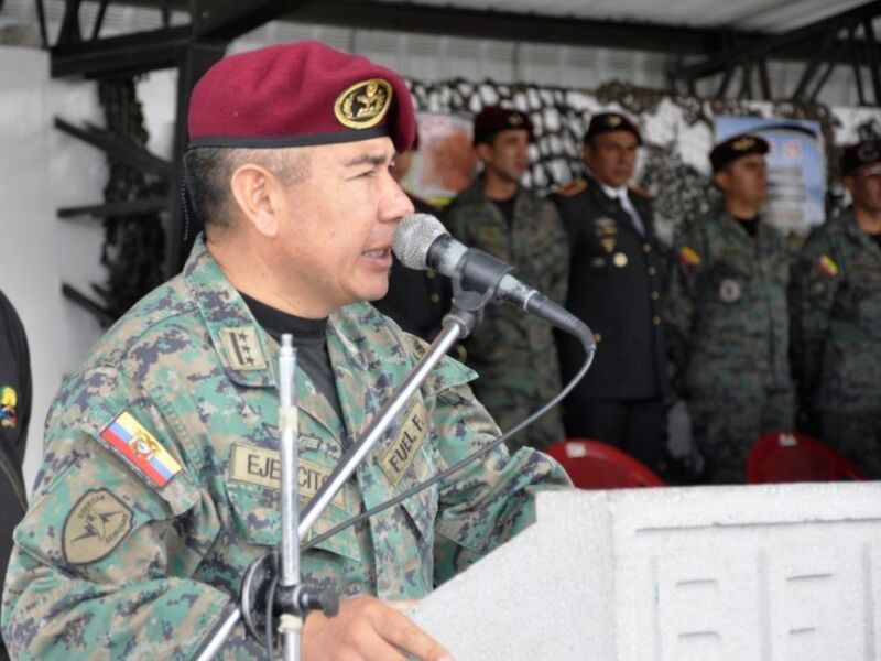 Un oficial militar habla en un evento público, rodeado por otros uniformados en un escenario al aire libre.