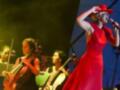 Una mujer en un vestido rojo con sombrero se levanta mientras canta, acompañada por músicos con violines y violonchelos en un escenario iluminado.